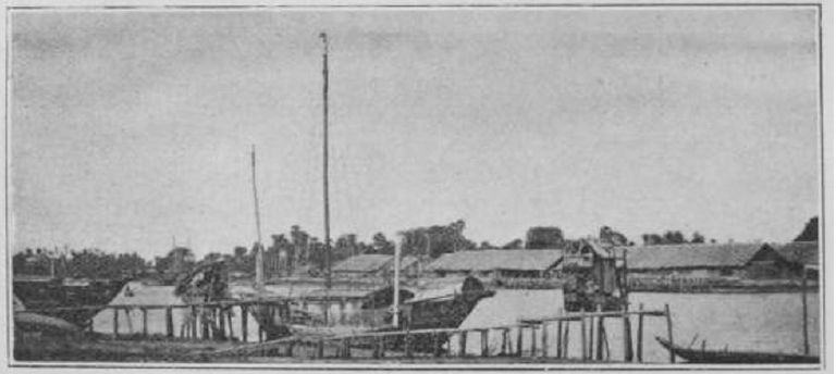 Boats moored along the Kampot river with low wooden buildings behind them in the 1880s